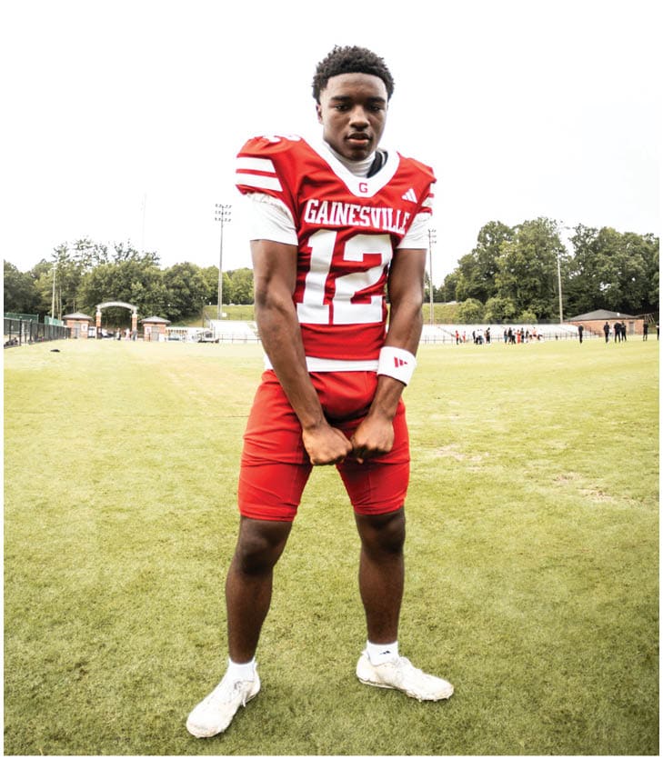A person wearing a red and white football uniform with the number 12 stands on a grassy field with trees and people in the background. Description generated by AI