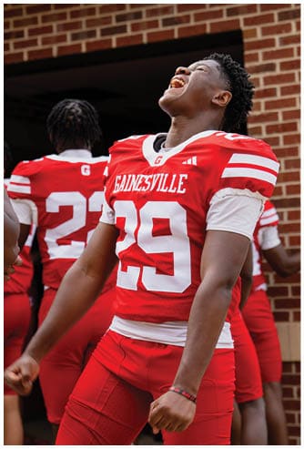 A person wearing a red and white Gainesville football jersey with the number 29 is standing with another person in a similar uniform visible in the background. Description generated by AI