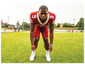 A person wearing a red football uniform with the number 9 is crouched in a ready position on a grassy field with a fence and trees in the background. Description generated by AI