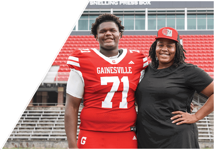 Two people stand side by side on a sports field, one wearing a red athletic uniform and the other wearing a black shirt and jeans. Description generated by AI