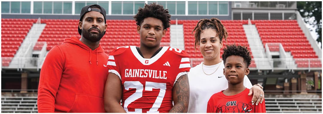 Four people stand together in a sports stadium with one person wearing a red football uniform and the others dressed casually. Description generated by AI