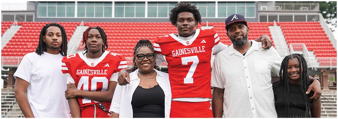 Five adults and one child stand together on a sports field with red bleachers in the background, two adults wearing red sports jerseys with “Gainesville" written on them. Description generated by AI
