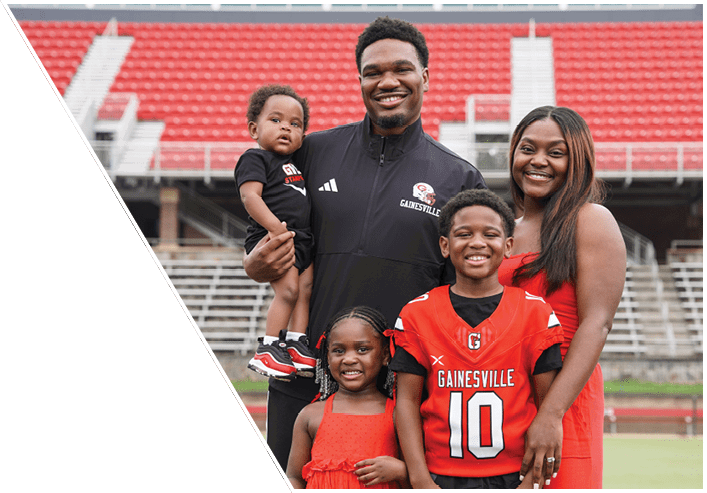 A group of five people, including two children wearing red Gainesville sports jerseys and three adults smiling together on a sports field with stadium seating in the background. Description generated by AI