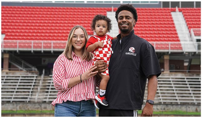 Two adults and a child stand in front of empty stadium seats with one adult holding the child who is wearing a red and white checkered outfit. Description generated by AI