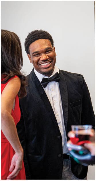 A person wearing a black velvet jacket and bow tie is smiling while standing next to two other people, one holding a glass of red wine. Description generated by AI