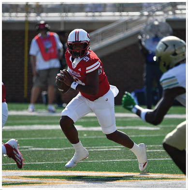 A person in a red and white football uniform is holding a football and preparing to throw while two other players, one in red and white and one in white and gold, are nearby on a football field. Description generated by AI