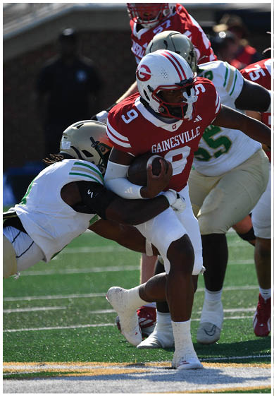 A person in a red and white football uniform is running with the ball while being tackled by a person in a white and green uniform on a football field. Description generated by AI