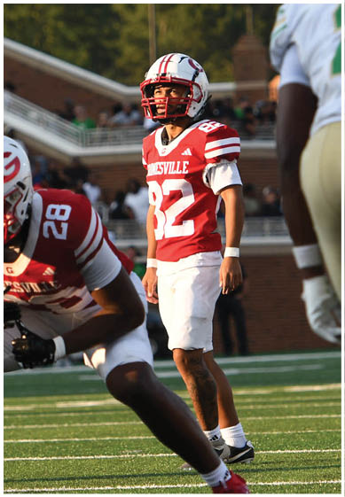 People in football uniforms are positioned on a field preparing for a play during a game with a crowd in the background. Description generated by AI