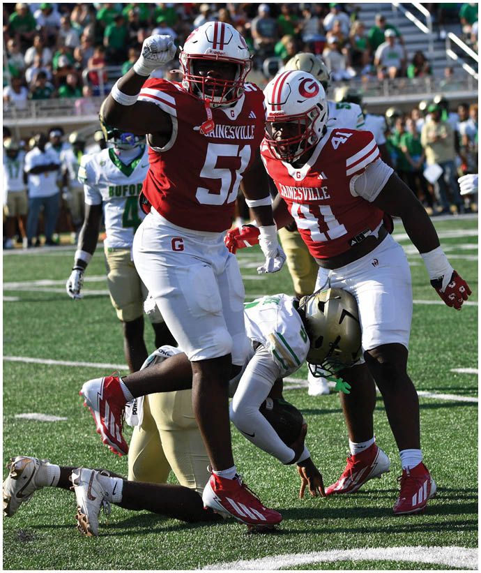Two football players in red and white uniforms celebrate on the field while a player in a white and green uniform is on the ground during a game. Description generated by AI