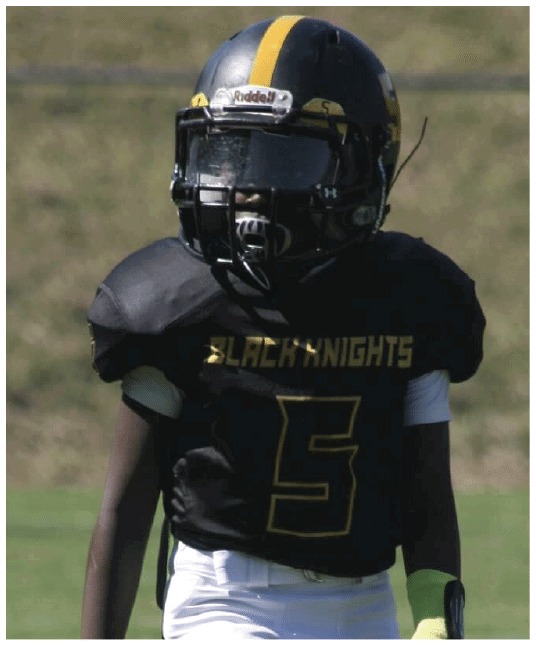A person wearing a black football uniform and a silver helmet with a yellow stripe is on a grassy field. Description generated by AI