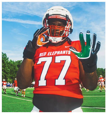 A person wearing a red football jersey with the number 77 and the words “RED ELEPHANTS" is standing on a football field making hand gestures while wearing a white helmet and gloves. Description generated by AI