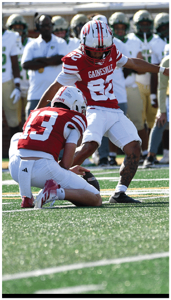 Two people in red and white football uniforms are on a field with one holding the ball in place while the other prepares to kick it. Description generated by AI