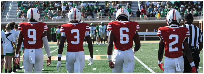Four people wearing red sports jerseys with white numbers and white pants stand side by side on a football field facing away from the camera. Description generated by AI
