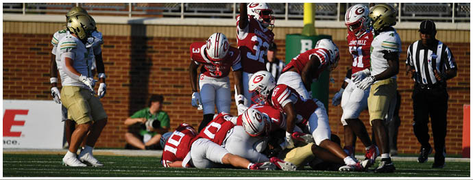 People in football uniforms are gathered on a field near a goalpost with one person raising an arm while a referee stands nearby. Description generated by AI