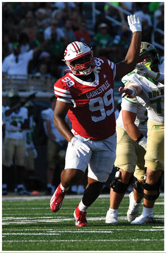 A person in a red football uniform with the number 99 is rushing towards a group of people in white and gold football uniforms, one of whom is holding a football. Description generated by AI