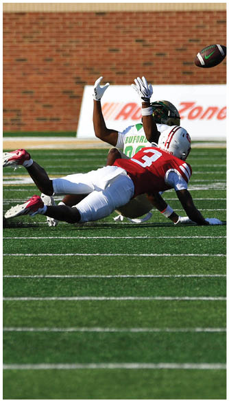 Two people in sports uniforms are on a football field with one diving and the other reaching up as a football flies above them. Description generated by AI