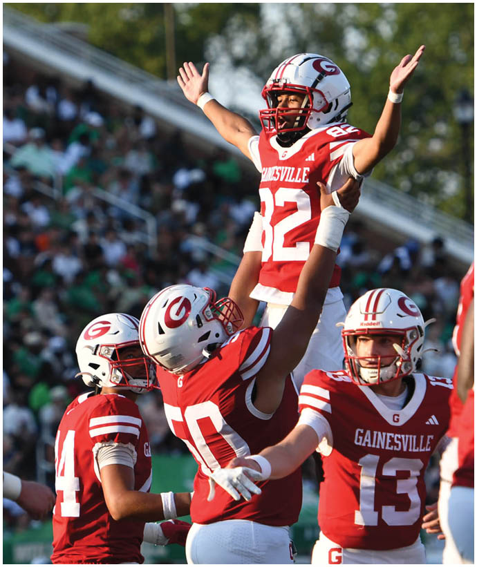 Four football players in red and white uniforms celebrate on the field with one player being lifted into the air while a crowd watches from the stands. Description generated by AI