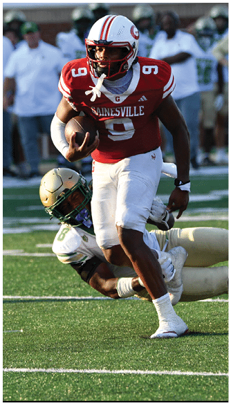 A person wearing a red football uniform with the number 9 is running with the ball while another person in a white and gold uniform attempts to tackle them on a football field. Description generated by AI