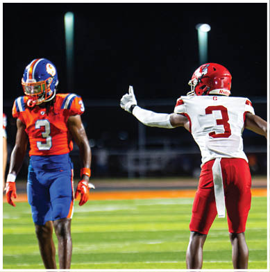 A person wearing a red football helmet and white jersey with the number 2 is giving a thumbs-up gesture on a dark field with another person in a blue and orange helmet partially visible. Description generated by AI