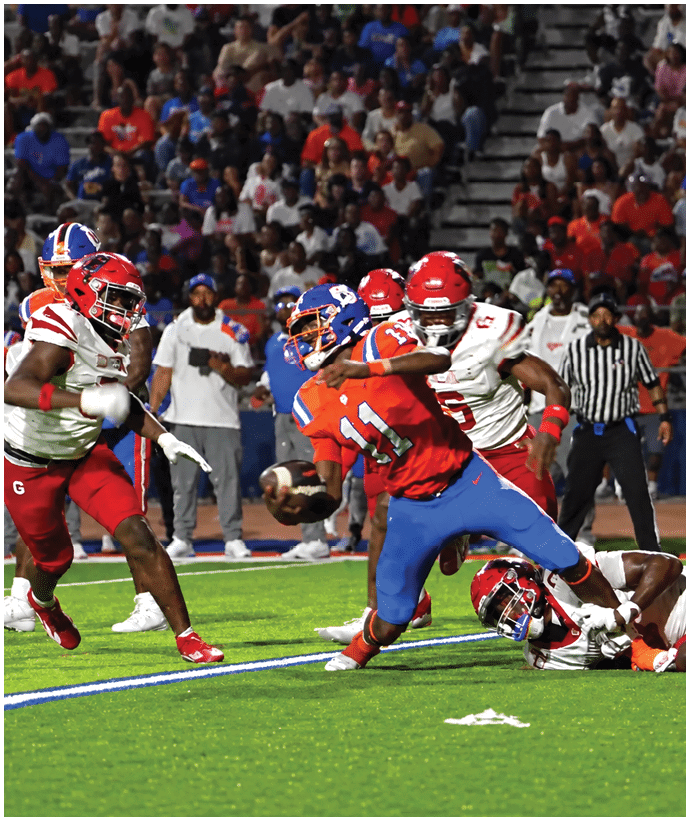 A person in a red and blue football uniform is running with the ball while being pursued and tackled by several people in white and red uniforms on a football field with a crowd in the background. Description generated by AI
