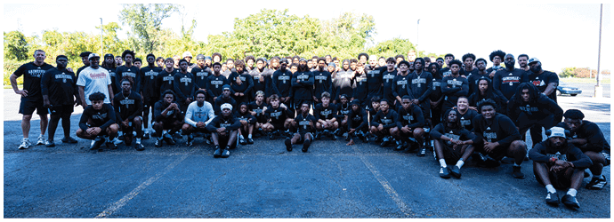 A large group of people wearing matching black shirts is gathered outdoors on a paved surface with trees and a clear sky in the background. Description generated by AI
