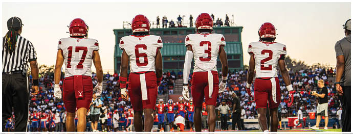 Four people in football uniforms and helmets stand side by side facing away from the camera with a scoreboard and spectators in the background. Description generated by AI