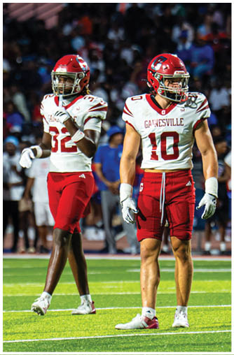 Two people wearing red and white football uniforms with helmets stand on a field with a crowd in the background during a game. Description generated by AI