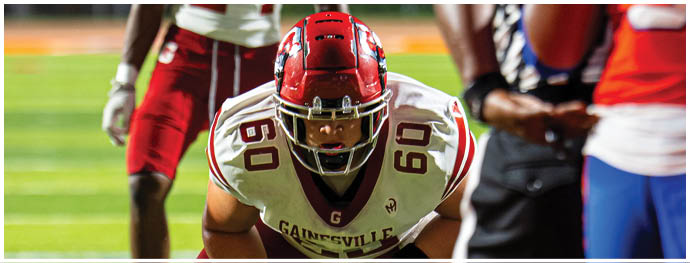 Two people in football uniforms are on a field, one wearing a white jersey with “Gainesville" and the number 24, and the other in an orange jersey with the number 3, with a referee partially visible between them. Description generated by AI