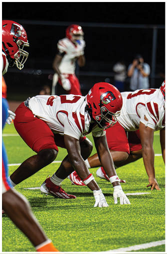 People in red and white football uniforms are crouched in a ready position on a green field during a nighttime game. Description generated by AI