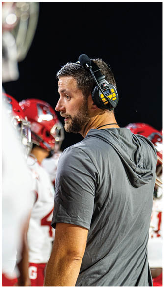 A person wearing a headset and a gray shirt stands on a sports field with a group of people in red and white football uniforms under bright stadium lights at night. Description generated by AI