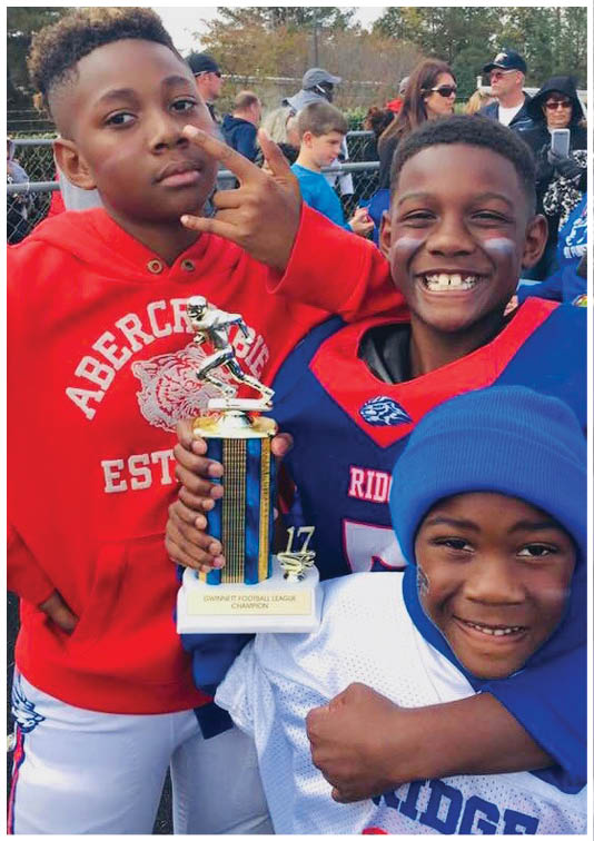 Three children wearing sports clothing smile and hold a football trophy outdoors with people and trees in the background. Description generated by AI