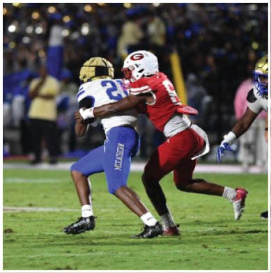 Two people in football uniforms are engaged in a tackle on a grassy field during a game with other players and spectators visible in the background. Description generated by AI