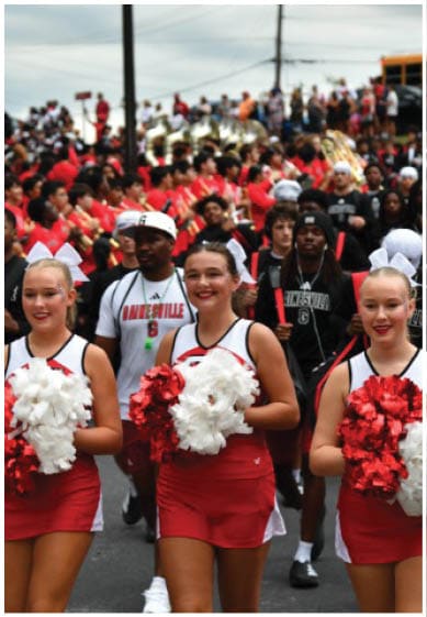 Two adults in red and white cheerleading uniforms hold red and white pom-poms while walking in front of a crowd of people, some wearing band uniforms and holding musical instruments. Description generated by AI