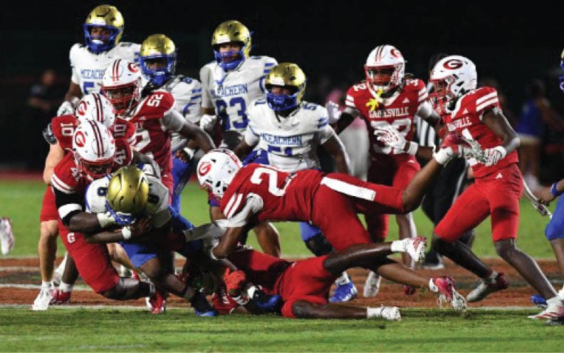 Several football players in red and white uniforms tackle a player in a blue and gold uniform on a grassy field during a nighttime game. Description generated by AI