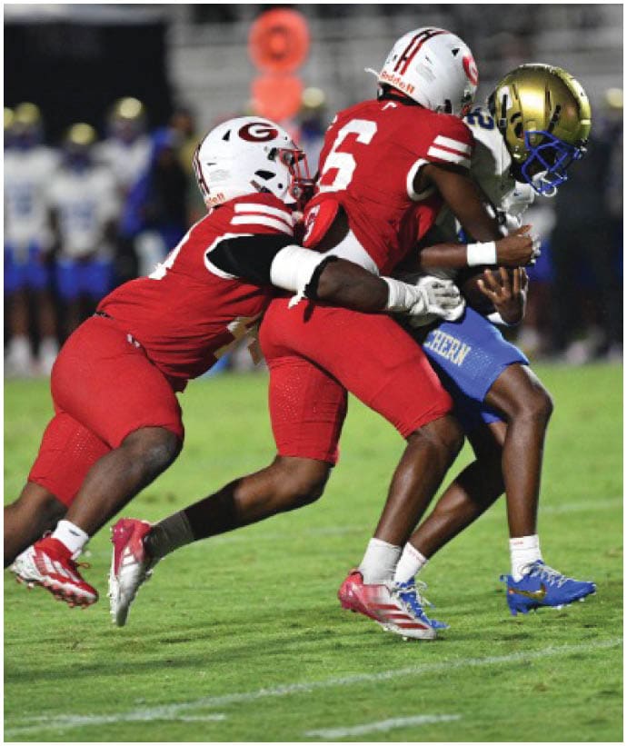 Two people in red football uniforms are engaged in a tackle during a game on a grass field with other players in blue uniforms in the background. Description generated by AI