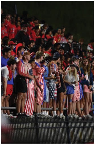People wearing various shorts and pants, including some with American flag patterns, are standing on a railing above a fence. Description generated by AI
