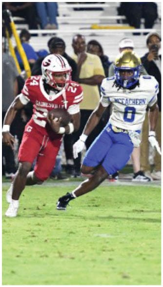 Two people in sports uniforms are running on a grass field during a football game, with one holding a football and the other attempting to catch or block. Description generated by AI