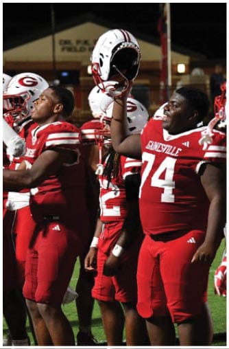 A group of people wearing red and white football uniforms are standing on a field at night, some holding their helmets up. Description generated by AI