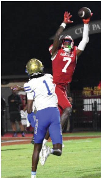 Two football players in white and blue uniforms are preparing to defend as a player in a red uniform jumps to catch or intercept the ball on a grassy field at night. Description generated by AI