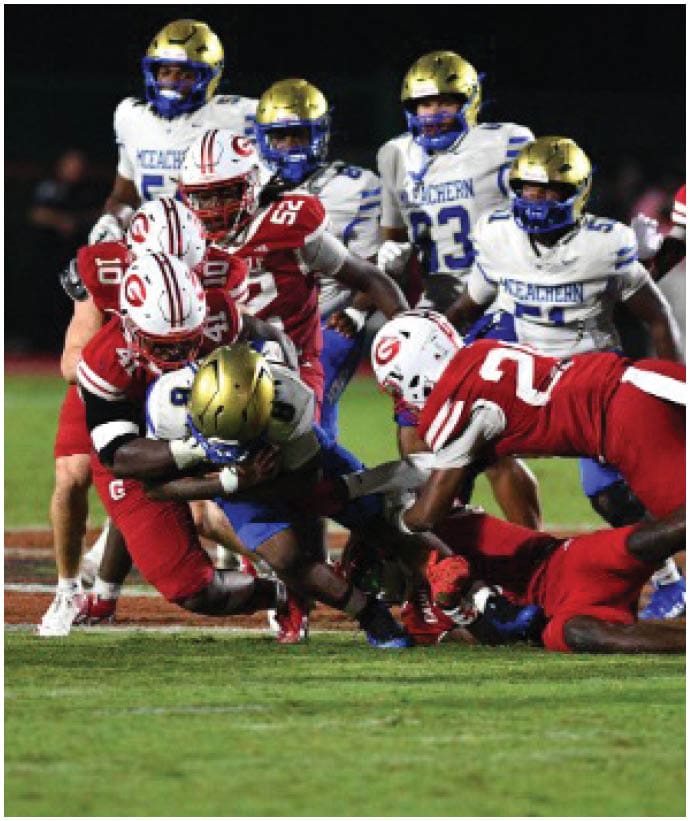 Players in red and white uniforms are tackling a player in a blue and gold uniform during a nighttime football game on a grass field. Description generated by AI