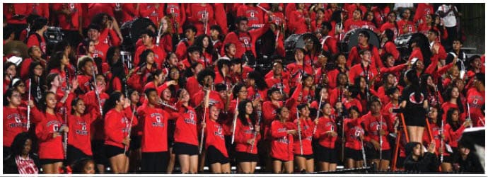 A large group of people wearing matching red shirts are standing closely together, some holding microphones and appearing to sing or cheer. Description generated by AI