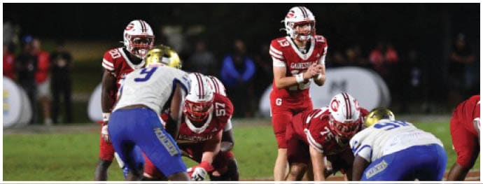 People in football uniforms are positioned on a field preparing for a play during a game at night. Description generated by AI