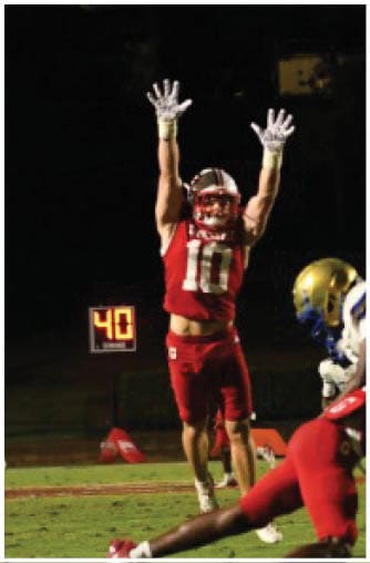 A person in a red football uniform is jumping with arms raised to catch a football while other players in red and blue uniforms are on the field during a nighttime game. Description generated by AI