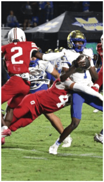 A person in a white and blue football uniform is holding a football while being tackled by a person in a red football uniform on a grassy field during a game. Description generated by AI