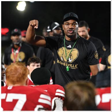 A person wearing a black Gainesville shirt and a whistle around their neck is gesturing with a raised arm while standing in front of people in red sports uniforms. Description generated by AI