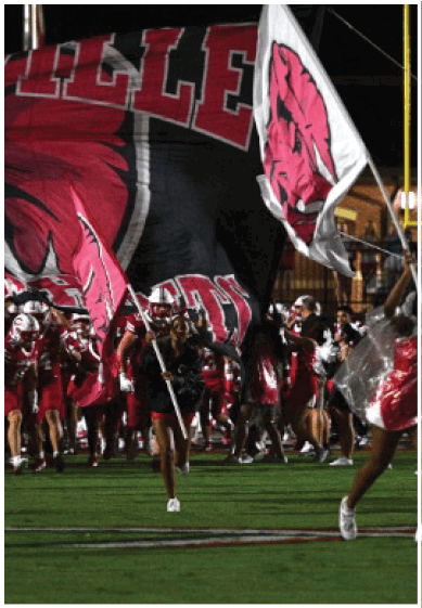 A group of football players in red and white uniforms run onto the field behind a person holding and waving a large flag with team colors and logo. Description generated by AI