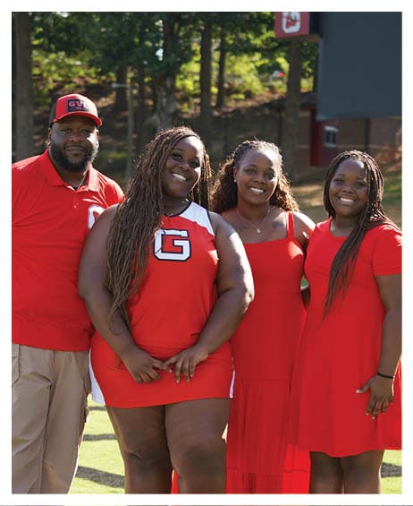 Two adults wearing red sports attire are standing outdoors on a grassy field with trees in the background, smiling and posing for the photo. Description generated by AI