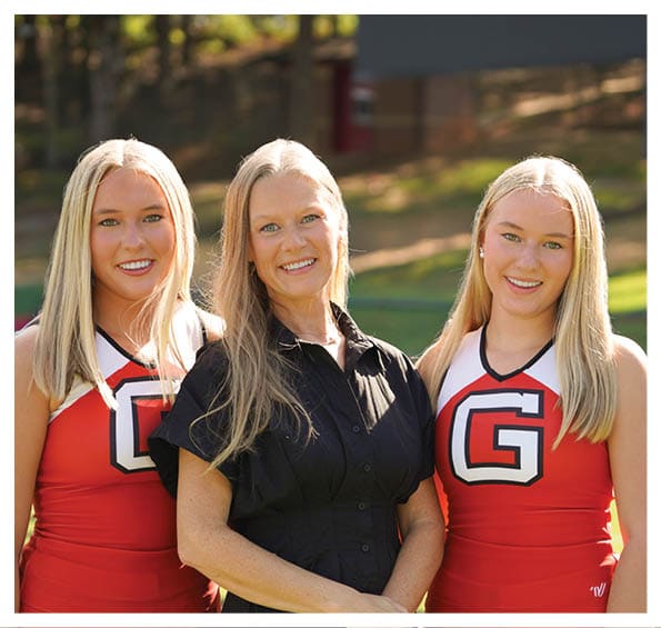 Three people are standing outdoors on grass with two wearing red cheerleading uniforms and one wearing a black dress, all smiling. Description generated by AI