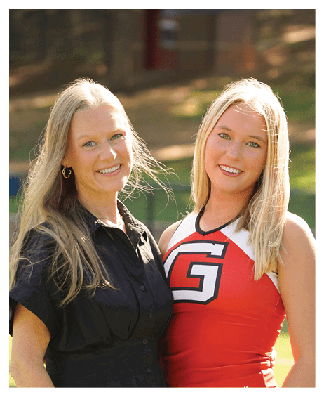 Two adults with long blonde hair are smiling outdoors with one wearing a black shirt and the other wearing a red and white cheerleading outfit. Description generated by AI