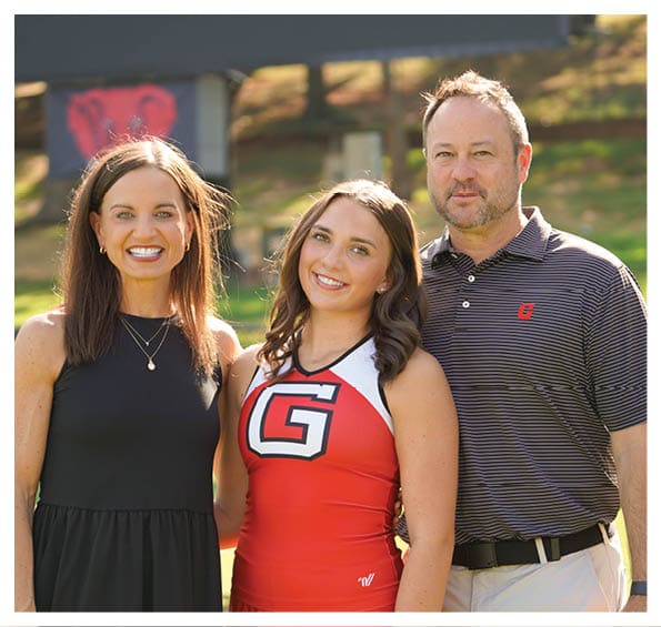 Three adults stand closely together outdoors with one wearing a red and white cheerleading uniform and the others dressed in casual clothing, all smiling. Description generated by AI